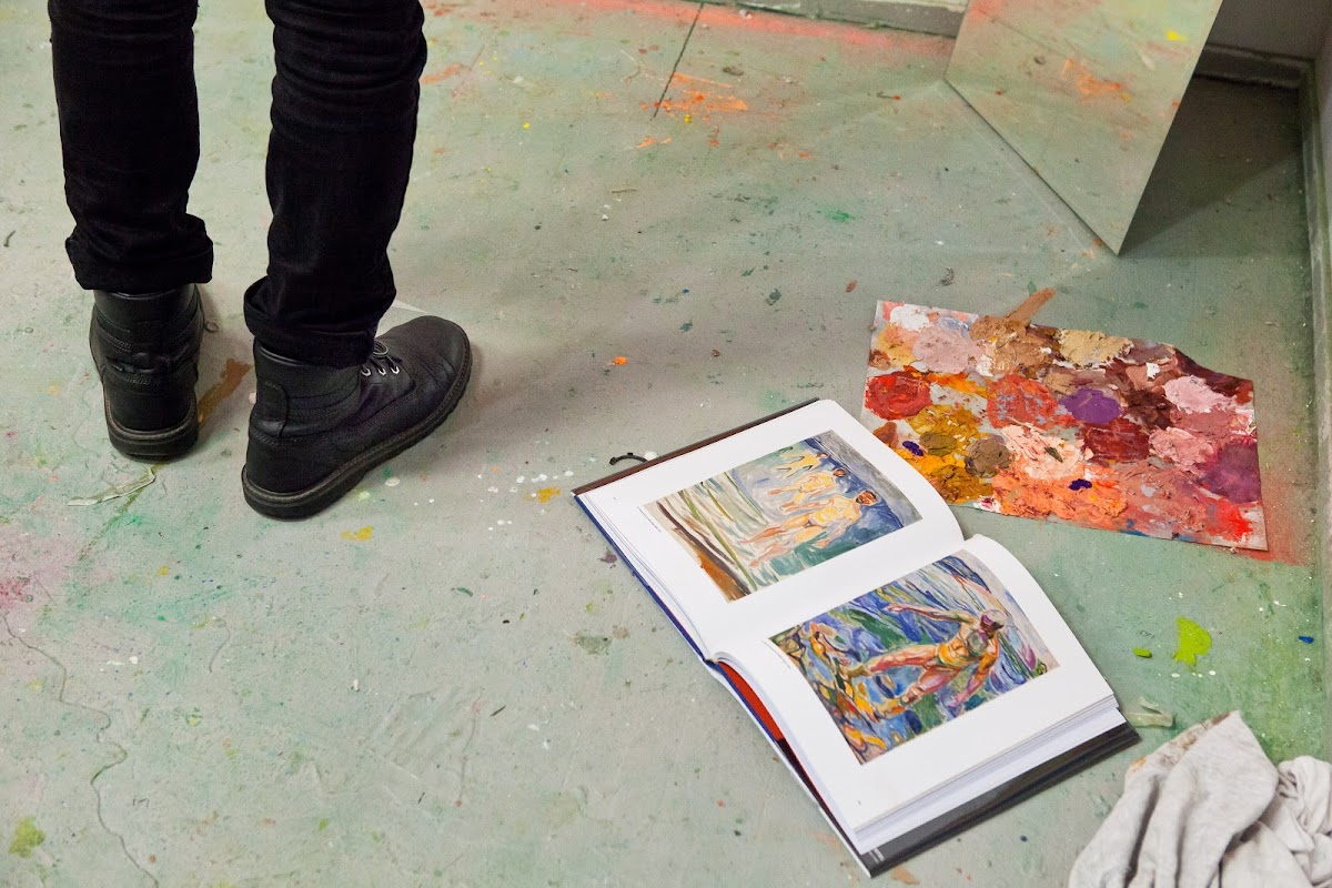 A person's feet standing in a studio with paint and an art book on the floor