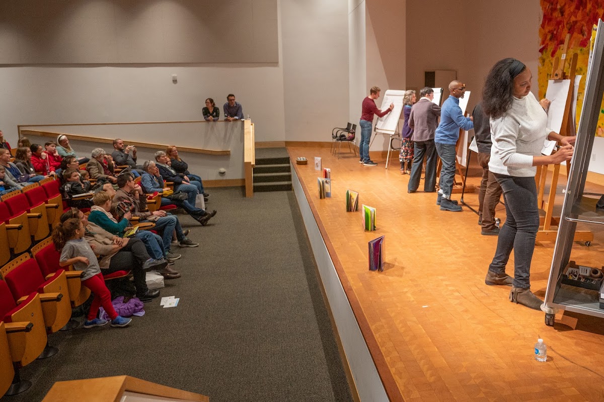 Artists draw at easels up on a stage as audience watches