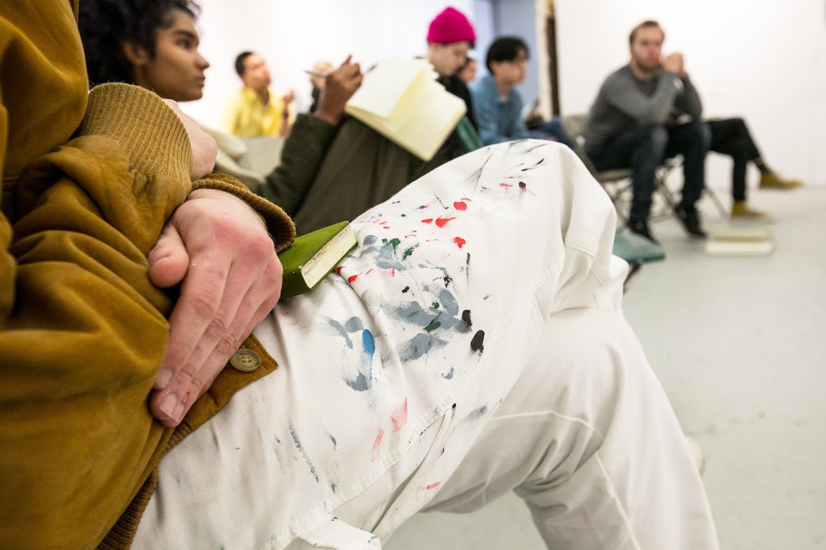 Close up of a student with paint-splattered pants sitting with other students during a critique