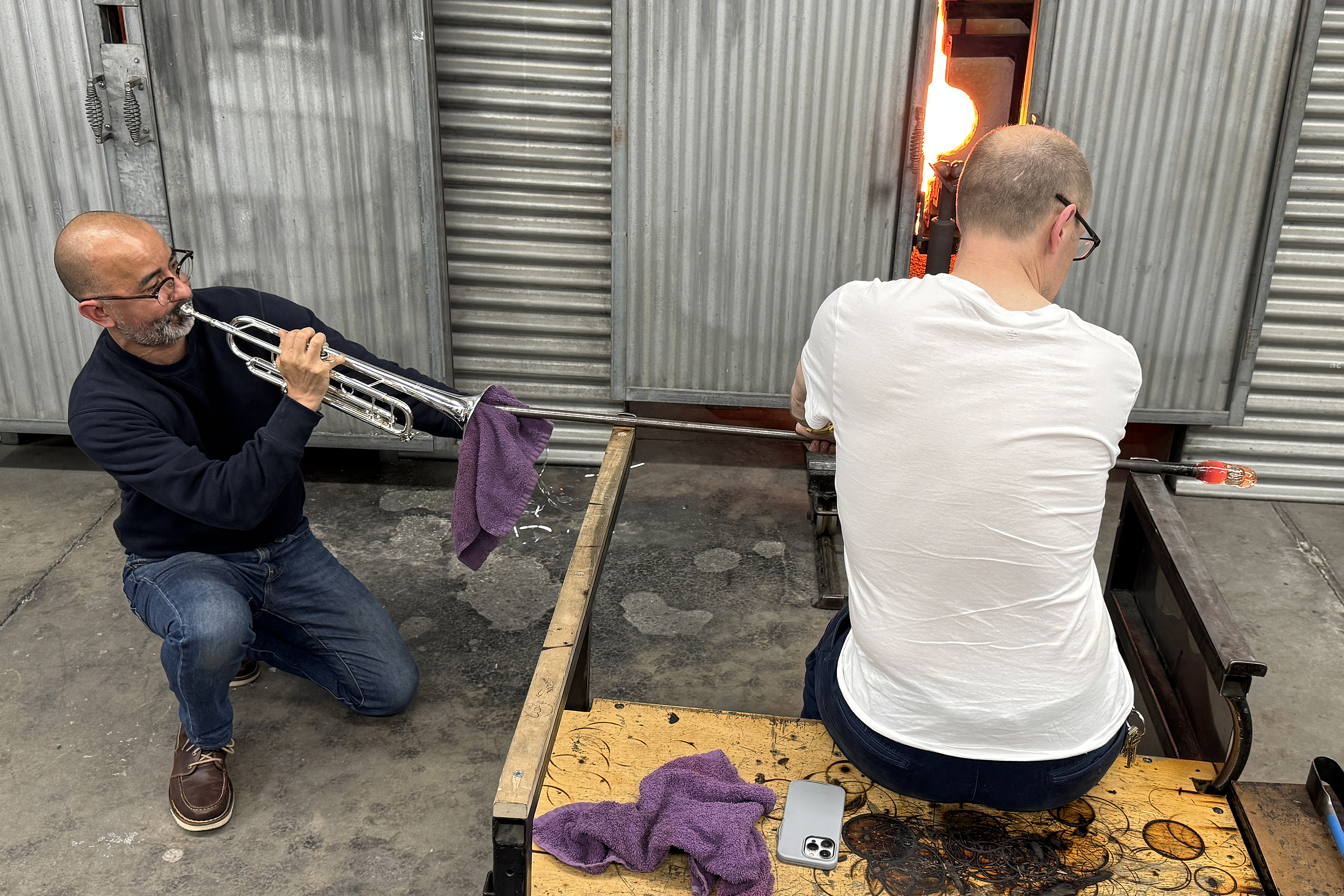 Valdez blows glass through his trumpet as Glass Department Head Sean Salstrom assists