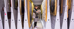 Student taking notes amongs stacks of paintings in a museum.