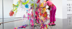 a student installs a piece suspending from the ceiling that is made up of colorful scraps of fabric