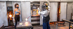 two glass students, one in protective gear, collaborate on a glassblowing piece