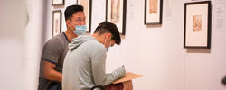 2 students sit and sketch in front of a row of artworks hanging in the risd museum