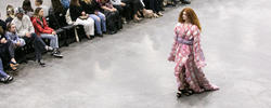 model with curly red hair walks down a runway in a pink and pillowy gown surrounded by audience members