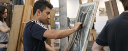 Student wearing a black t-shirt in a drawing studio sketching at an easel with charcoals