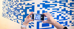 two hands hold a smartphone in front of a large-scale, blue-on-white mural of a QR code