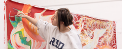 Student painting on a hanging canvas fabric using bright bold reds, pinks, golds and greens