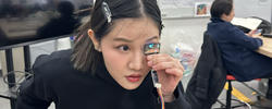 a student looks through a lens attached to several wires, in a classroom setting with a person at a desk in the background