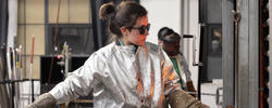 student wearing silver protective coat and protective glasses works with a boro rod inside a glass studio