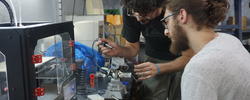 two people look closely as they work on a robotic object at a workstation equipped with other technological objects