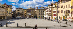 a city square in Florence, Italy, with people moving on foot or bike, under a blue sky with white clouds