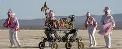 four people walk alongside a mechanical horse frame in the desert, with a mountain range and blue sky far in the distance