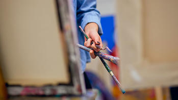 person painting on an easel while holding tools in their opposite hand