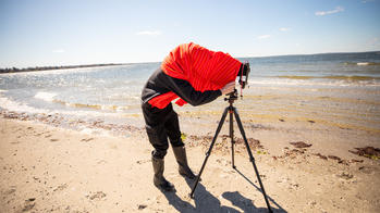 an R I S D student covers their head with a red jacket while photographing on a sandy beach along the coastline