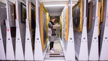 Student taking notes amongs stacks of paintings in a museum.