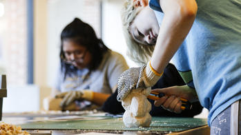 students at a worktable use hand tools to shape wood into sculptures