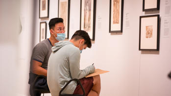 2 students sit and sketch in front of a row of artworks hanging in the risd museum