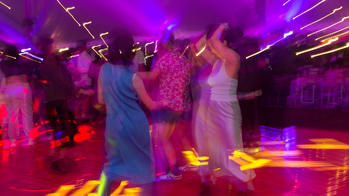 a dancer in a blue dress and another in a silver and white outfit dance on a red-lit dance floor (with purple-lit ceiling above) with several other blurred figures in the background