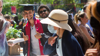 a student in graduation regalia looks at camera as others surround him in foreground and background at an outdoor party