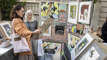 a person in a pale orange coat admires an artwork as the artist looks on while surrounded by other paintings and drawings at an outdoor vendor booth