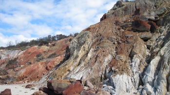 a cliff face of red, brown and gray rock running along a sandy beach, with slightly cloudy blue sky in the background