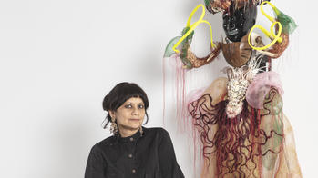 Rina Banerjee sitting next to a sculpture of a humanoid figure with a black head and gritted white teeth, wearing a dress decorated with shells, potatoes and lacy fabrics