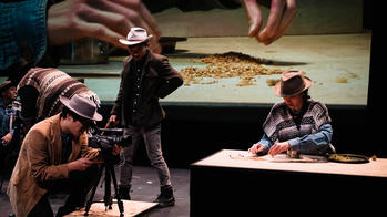 five people dressed in Western-style coats and hats work on a film set as a screen in the background projects a pair of hands working with a grain-like substance