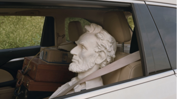 a stone Abe Lincoln head strapped into the driver's seat of a car