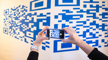 two hands hold a smartphone in front of a large-scale, blue-on-white mural of a QR code