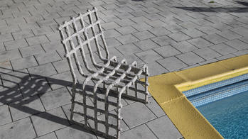 a grayish white chair, built with rough, pole-like parts in a grid, sits by the corner of an in-ground pool