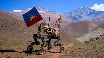 a robotic mule carries a blue, red and gold flag through mountain territory toward blue skies