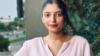 Areeha Ahmad in a pink cotton v-neck top hair pulled back in front of a wall with vines and a city in the distance