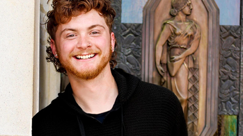 Jeremy Grail in a black tshirt standing against a wall in front of a wood carving