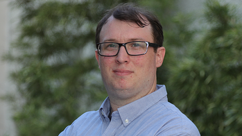 Matthew Brown in a light blue button down shirt wearing glasses in front of a wall with vines