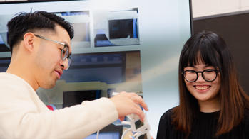 two students look at a pair of glasses with square black lenses, with a large monitor in the background