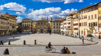 a city square in Florence, Italy, with people moving on foot or bike, under a blue sky with white clouds