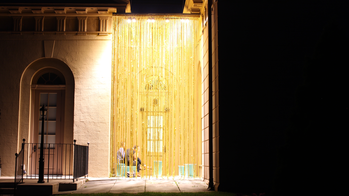 people sit behind a large yellow-beaded curtain hanging illuminated from the roof of a 19th-century style building