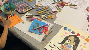 a child’s hand holds a pencil while working at a drawing table covered by art papers and supplies