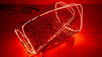 a thin strip of red neon lighting wrapped around repurposed bed springs in a rectangular cube formation, inside a red-lit room