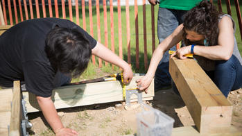 two people build a small wood structure within the frame of an outdoor sandbox
