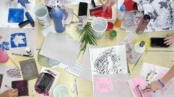 Table covered in art supplies with various students reaching across it to work on their art projects through project open door.