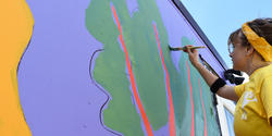Graduate Student Simone Nemes stands on a ladder while painting stalks of kale on a shipping container at ECO City Farms