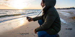 a student wearing a parka squats in front of the breaking waves to record