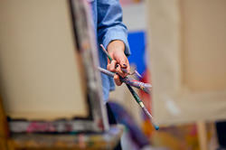 person painting on an easel while holding tools in their opposite hand