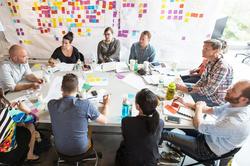 RISD faculty and others sit around a table, in front of an idea board filled with multicolored post-its