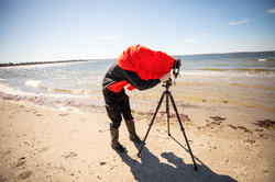 an R I S D student covers their head with a red jacket while photographing on a sandy beach along the coastline