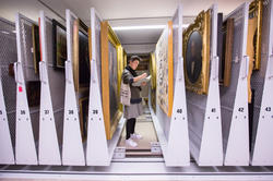 Student taking notes amongs stacks of paintings in a museum.