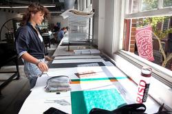 a student works in a RISD studio with a RISD flag flying outdoors, visible through a window