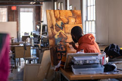 Illustration students sits in a bright studio space painting, surrounded by books, a palette, and paints
